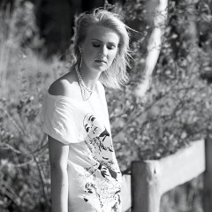 A young woman with short fair hair sits by a wooden fence outdoors, wearing a light T-shirt printed with graphics, looking down with her eyes closed.